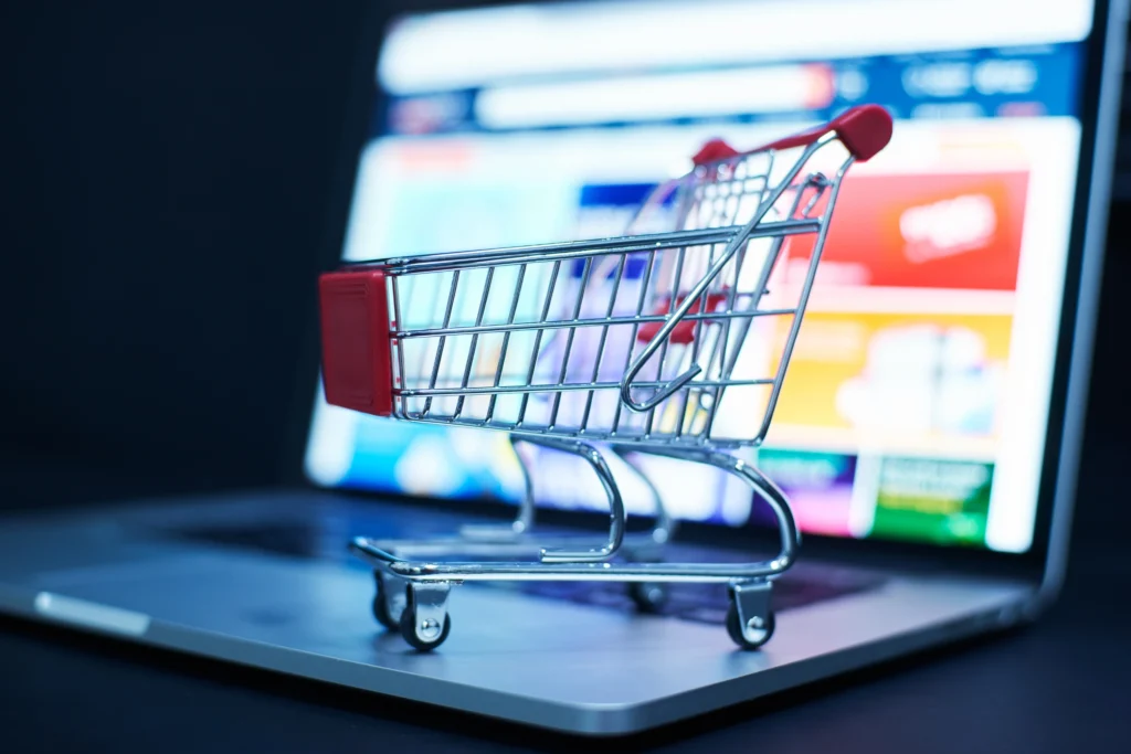 Un pequeño carrito de compras de supermercado en miniatura apoyado sobre un portátil abierto, con la pantalla del ordenador mostrando una página web de compras online de fondo borroso. Simboliza el comercio electrónico y la acción de añadir productos al carrito.