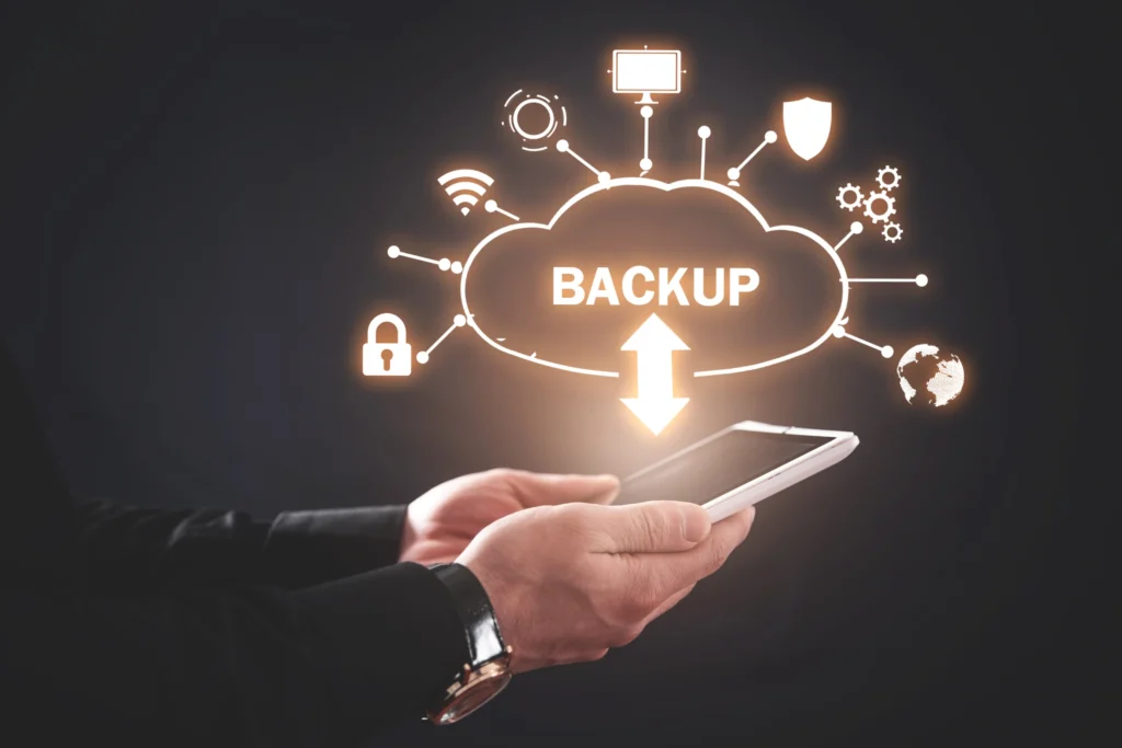 Manos sosteniendo un smartphone, con un icono de nube brillante que dice 'BACKUP' en el centro, conectado a varios iconos de seguridad y datos. Simboliza la importancia de las copias de seguridad en la nube para un negocio online.