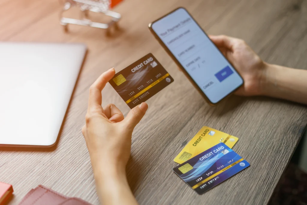 Persona sosteniendo una tarjeta de crédito frente a un teléfono móvil con un formulario de pago, con más tarjetas sobre una mesa, simbolizando métodos de pago y transacciones online.