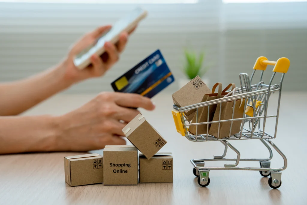 Manos sosteniendo un móvil y una tarjeta de crédito, junto a cajas de cartón con la etiqueta "Shopping Online" y un pequeño carrito de supermercado lleno de paquetes, simbolizando una compra y envío en un marketplace.