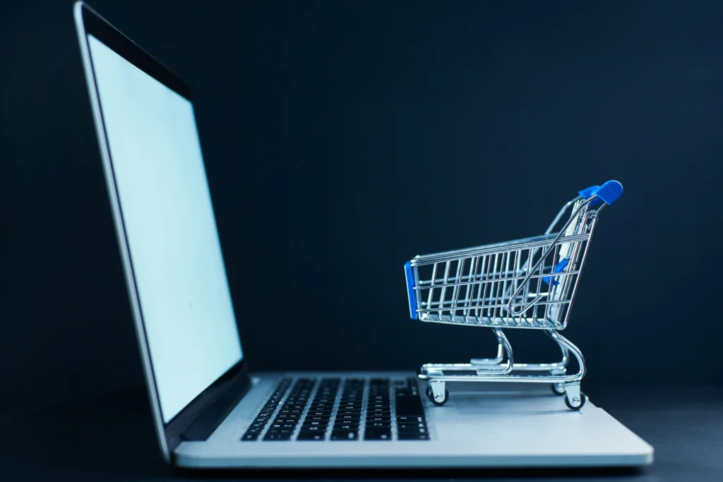 Una computadora portátil abierta con un pequeño carrito de compras de supermercado en miniatura sobre su teclado, simbolizando el comercio electrónico y las compras online.