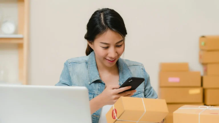 Emprendedora revisa un pedido con su teléfono junto a su laptop y cajas listas para enviar